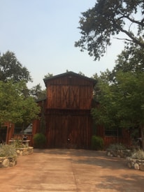 A rustic wooden barn surrounded by lush greenery and trees. The structure has large double doors and a prominent triangular roof. There are neatly arranged stone planters with shrubs on either side of the entrance. The setting appears calm and serene, situated on a paved area.
