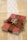 A small red book is wrapped with a brown burlap ribbon, which is tied into a bow on top. Two intertwined gold rings are placed on the bow. The book rests on a burlap cloth with a decorative white lace strip running vertically underneath.