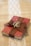A small red book is wrapped with a brown burlap ribbon, which is tied into a bow on top. Two intertwined gold rings are placed on the bow. The book rests on a burlap cloth with a decorative white lace strip running vertically underneath.