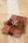 A small red book is wrapped with a brown burlap ribbon, which is tied into a bow on top. Two intertwined gold rings are placed on the bow. The book rests on a burlap cloth with a decorative white lace strip running vertically underneath.