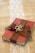 A small red book is wrapped with a brown burlap ribbon, which is tied into a bow on top. Two intertwined gold rings are placed on the bow. The book rests on a burlap cloth with a decorative white lace strip running vertically underneath.