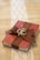 A small red book is wrapped with a brown burlap ribbon, which is tied into a bow on top. Two intertwined gold rings are placed on the bow. The book rests on a burlap cloth with a decorative white lace strip running vertically underneath.