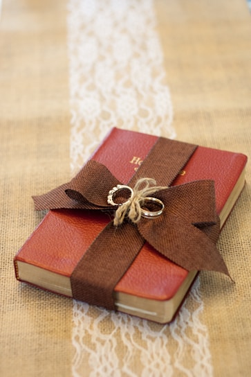 A small red book is wrapped with a brown burlap ribbon, which is tied into a bow on top. Two intertwined gold rings are placed on the bow. The book rests on a burlap cloth with a decorative white lace strip running vertically underneath.