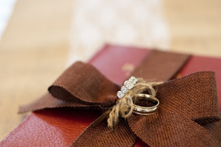 A brown fabric ribbon tied in a bow adorns the top of a reddish-brown textured surface, possibly a gift box. Two rings rest on the bow, one featuring a row of sparkling gemstones. The background is softly blurred, highlighting the rings and bow.