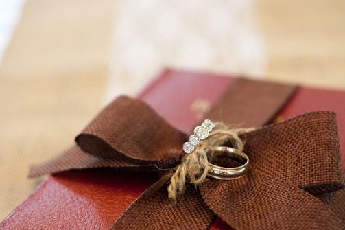 A brown fabric ribbon tied in a bow adorns the top of a reddish-brown textured surface, possibly a gift box. Two rings rest on the bow, one featuring a row of sparkling gemstones. The background is softly blurred, highlighting the rings and bow.