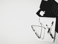 An overhead view of a black keyboard and a notepad resting on a white surface. A pair of eyeglasses and a pen are placed on the notepad. The Apple logo is visible on one of the items, possibly indicating a laptop or tablet.