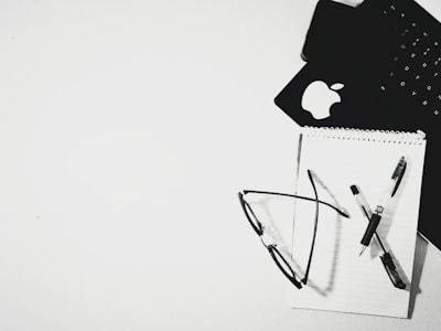 An overhead view of a black keyboard and a notepad resting on a white surface. A pair of eyeglasses and a pen are placed on the notepad. The Apple logo is visible on one of the items, possibly indicating a laptop or tablet.