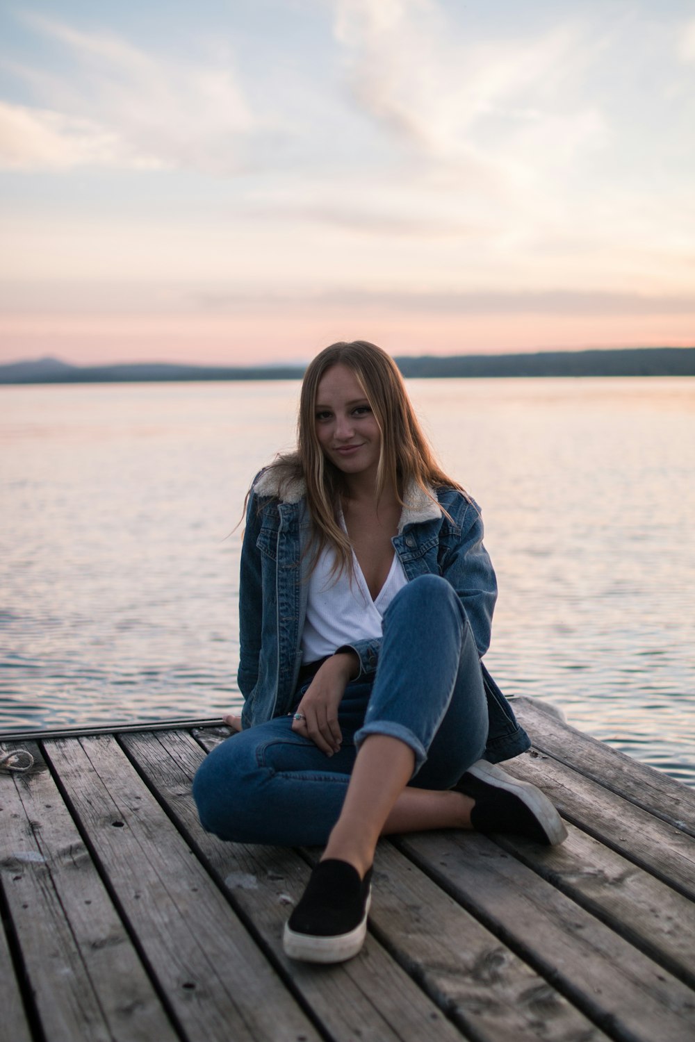 woman in white v-neck shirt wearing blue denim jacket sitting on brown wooden dock beside ocean water during daytime