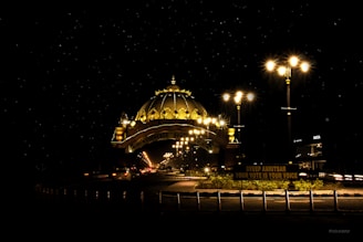 A beautifully lit dome structure illuminates the night with its golden glow. Streetlights line the road, adding to the bright atmosphere as vehicles pass by. A sign at the forefront highlights a civic message about voting.