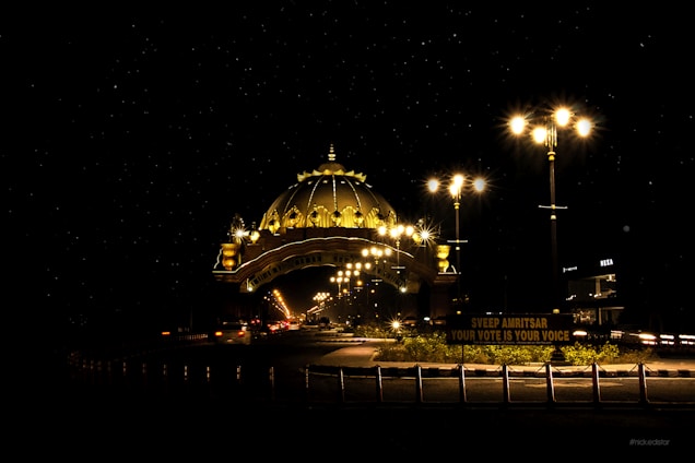 A beautifully lit dome structure illuminates the night with its golden glow. Streetlights line the road, adding to the bright atmosphere as vehicles pass by. A sign at the forefront highlights a civic message about voting.