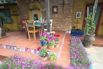 An inviting outdoor patio features terracotta tiles, a rustic wooden table, and chairs. Clay pots bursting with vibrant flowers sit on steps decorated with colorful mosaic patterns. The backdrop includes a stone wall with decorative lanterns and a hanging iron cross. A large potted plant adds greenery, while a sign reading 'Soul Siesta' hangs next to a religious picture.