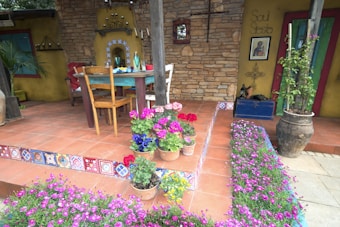 An inviting outdoor patio features terracotta tiles, a rustic wooden table, and chairs. Clay pots bursting with vibrant flowers sit on steps decorated with colorful mosaic patterns. The backdrop includes a stone wall with decorative lanterns and a hanging iron cross. A large potted plant adds greenery, while a sign reading 'Soul Siesta' hangs next to a religious picture.