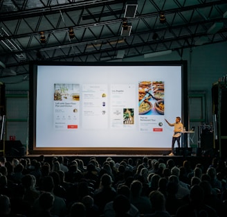 person discussing while standing in front of a large screen in front of people inside dim-lighted room