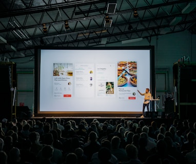 person discussing while standing in front of a large screen in front of people inside dim-lighted room