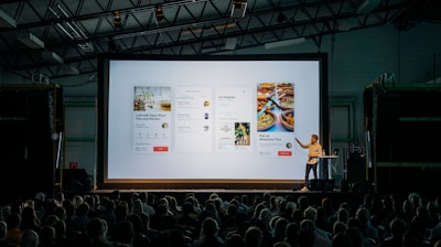 person discussing while standing in front of a large screen in front of people inside dim-lighted room