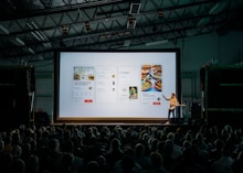 person discussing while standing in front of a large screen in front of people inside dim-lighted room