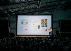 person discussing while standing in front of a large screen in front of people inside dim-lighted room