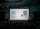 person discussing while standing in front of a large screen in front of people inside dim-lighted room