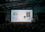 person discussing while standing in front of a large screen in front of people inside dim-lighted room