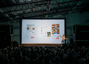 person discussing while standing in front of a large screen in front of people inside dim-lighted room