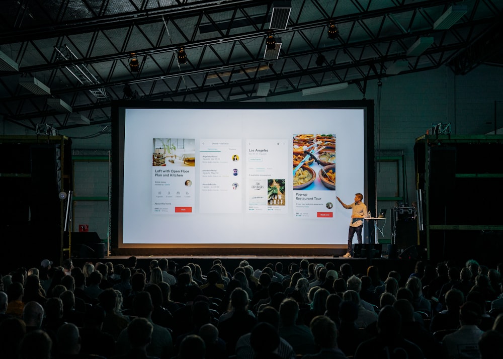 person discussing while standing in front of a large screen in front of people inside dim-lighted room