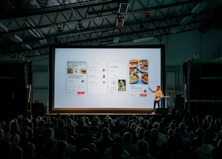 person discussing while standing in front of a large screen in front of people inside dim-lighted room