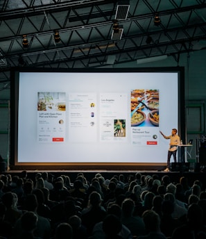 person discussing while standing in front of a large screen in front of people inside dim-lighted room