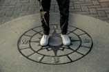 A person stands on a compass rose embedded in pavement, with cardinal directions marked as N, E, S, W. The person wears white sneakers and green pants, and the scene features a combination of stone and brick surfaces.