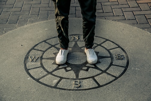A person stands on a compass rose embedded in pavement, with cardinal directions marked as N, E, S, W. The person wears white sneakers and green pants, and the scene features a combination of stone and brick surfaces.
