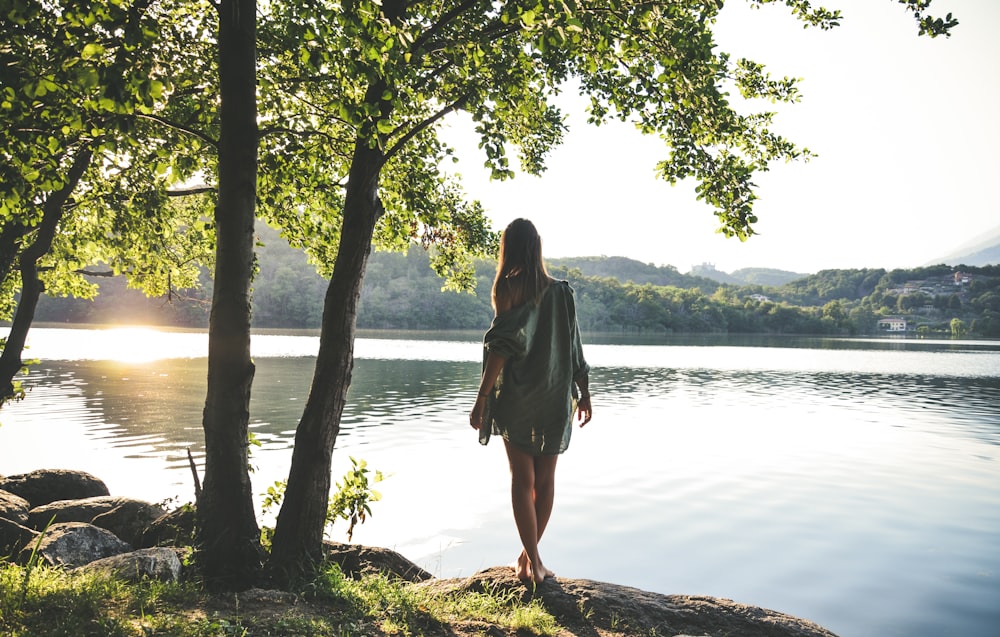 woman standing beside body of water and trees near mountains under white sky during daytime