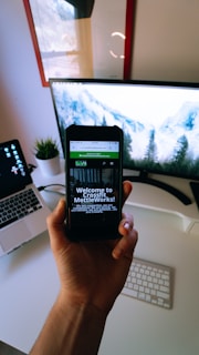 A person is holding a smartphone displaying a website with the text 'Welcome to Crossfit MettleWorks!'. The background shows a modern workspace setup with a desktop computer displaying a scenic mountain image, a laptop, a small potted plant, a wireless keyboard, and a reflective, framed picture on the wall.