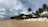A tranquil beach scene with rows of white lounge chairs facing the ocean. Palm trees are scattered along the edge of the sandy shore. A series of white canopies provide shade. The sky is partly cloudy, with a hint of overcast in the distance.