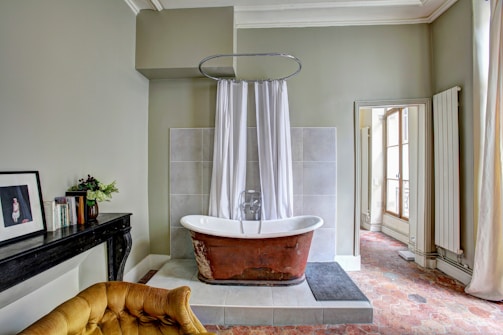A vintage-style bathroom featuring a freestanding rust-colored bathtub with white inner lining, surrounded by a curtain on a circular rod. The space is decorated with pale green walls and a gray tiled accent wall behind the tub. To the left, there is a dark wooden table with a framed painting, books, and a small vase with flowers. A plush mustard-colored chair is partially visible at the bottom of the image. Sunlight is streaming in through a large, partially curtained window on the right, illuminating a patterned floor.