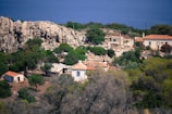 A rural landscape with stone houses featuring red-tiled roofs nestled among rocky terrain and lush greenery. In the background, a calm, deep blue sea stretches across the horizon, while a rugged cliffside looms to the left.