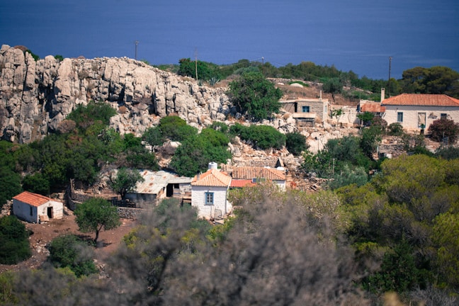 A rural landscape with stone houses featuring red-tiled roofs nestled among rocky terrain and lush greenery. In the background, a calm, deep blue sea stretches across the horizon, while a rugged cliffside looms to the left.