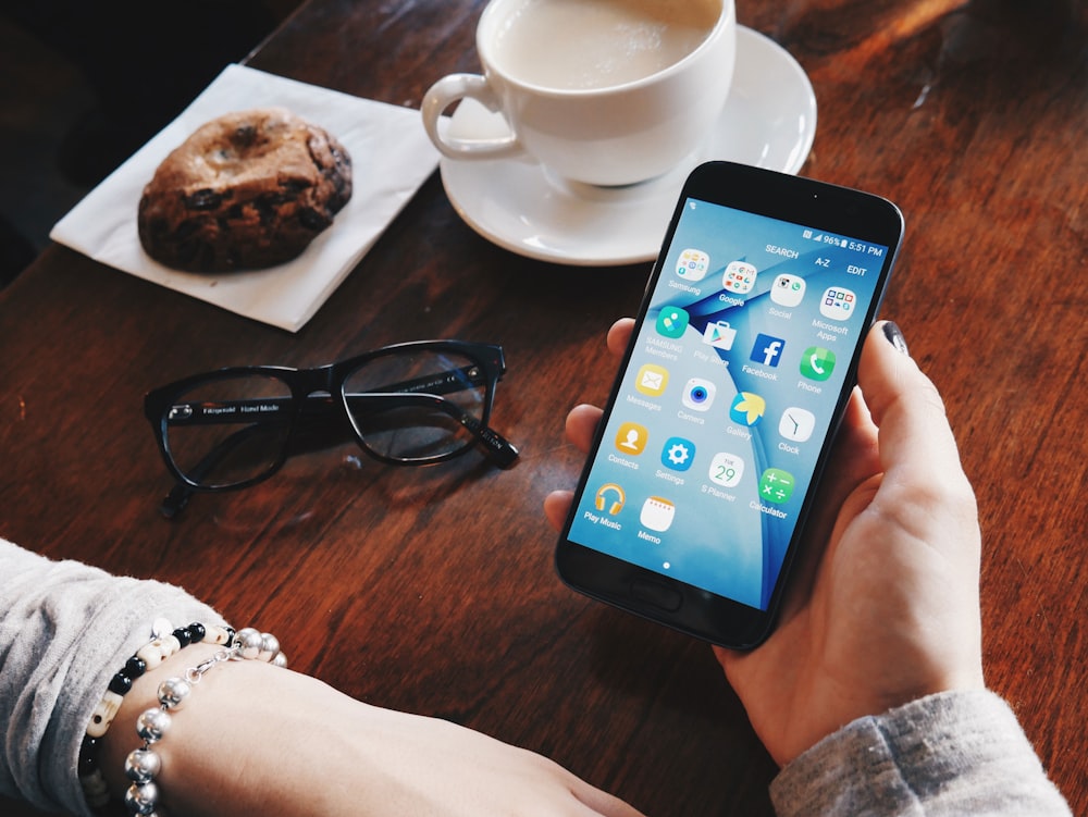 Person mit eingeschaltetem Smartphone sitzt am Tisch mit gefüllter Tasse mit Untertasse