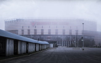 A large, modern arena with a striped exterior and prominent signage partially obscured by fog. The foreground features an empty, overcast area with a few street lamps and a structure along one side.