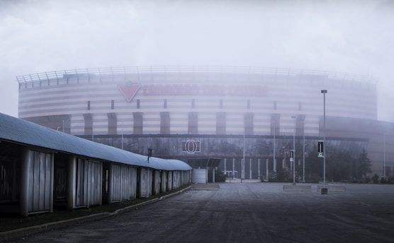 A large, modern arena with a striped exterior and prominent signage partially obscured by fog. The foreground features an empty, overcast area with a few street lamps and a structure along one side.