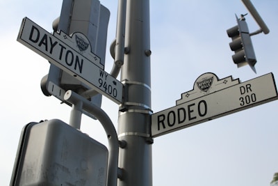 Street signs at a junction showing Dayton Way and Rodeo Drive with a Beverly Hills emblem, mounted on a gray metal pole. A traffic light and power box are visible against a cloudy sky.