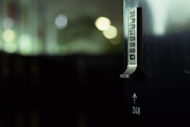 A metallic lock with a numeric keypad is mounted on a dark surface, with blurred lights and shadows in the background. The image highlights the lock's sleek design and the numbers displayed on the keypad.