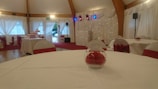 An elegantly decorated event hall with round tables covered in white tablecloths and red accents, such as chair bows and floral table centerpieces. The room has draped curtains and wooden beams, with stage lighting in the background.