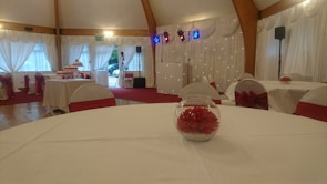 An elegantly decorated event hall with round tables covered in white tablecloths and red accents, such as chair bows and floral table centerpieces. The room has draped curtains and wooden beams, with stage lighting in the background.