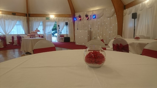 An elegantly decorated event hall with round tables covered in white tablecloths and red accents, such as chair bows and floral table centerpieces. The room has draped curtains and wooden beams, with stage lighting in the background.