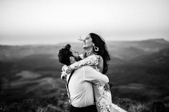 A joyful couple is captured in an embrace with a scenic mountainous backdrop. The woman is laughing, and her hair flows in the wind, while the man holds her tightly with a broad smile. She is wearing a delicate lace dress, and the scene is presented in black and white, adding a timeless feel.