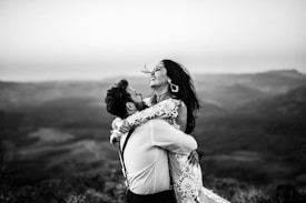 A joyful couple is captured in an embrace with a scenic mountainous backdrop. The woman is laughing, and her hair flows in the wind, while the man holds her tightly with a broad smile. She is wearing a delicate lace dress, and the scene is presented in black and white, adding a timeless feel.