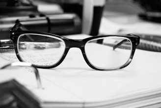 A pair of eyeglasses with dark frames rests on an open notebook, surrounded by various office supplies such as pens and papers.