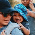 A smiling baby wears a colorful hat with a pattern of dinosaurs, sitting on an adult's shoulder. The adult, wearing sunglasses and a black cap, looks ahead. In the background, another person appears slightly blurred.