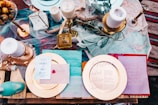 An elegantly set table featuring a variety of decorative items including candles, a glass lantern, and an assortment of plates with menus. The arrangement includes textured fabrics and foliage, creating a rustic yet sophisticated ambiance. Gold and pastel hues are prominent, with elements like a red wax seal adding a touch of classic charm.