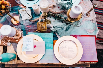 An elegantly set table featuring a variety of decorative items including candles, a glass lantern, and an assortment of plates with menus. The arrangement includes textured fabrics and foliage, creating a rustic yet sophisticated ambiance. Gold and pastel hues are prominent, with elements like a red wax seal adding a touch of classic charm.
