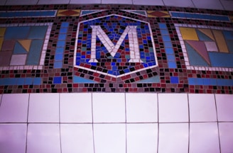 A colorful mosaic featuring an arrangement of small, square tiles forming an abstract design. At the center, the letter 'M' stands out prominently, surrounded by geometric patterns in various hues. The background consists of larger square tiles.